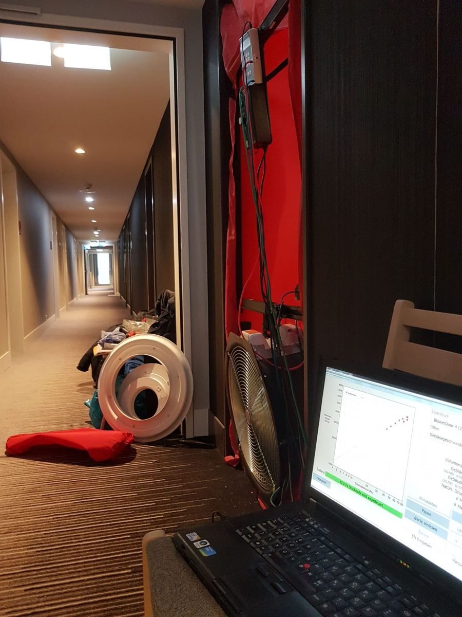Blower-Door-Messgerät mit Gebläse und Messrahmen in einer Tür, daneben ein Laptop mit der Messkurve, in einem Hotelflur